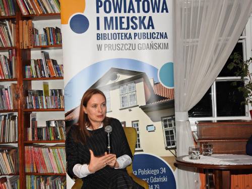 Autorka Katarzyna Pełczyńska-Nałęcz podczas spotkania w bibliotece. Kobieta siedzi n tle plakatu biblioteki.